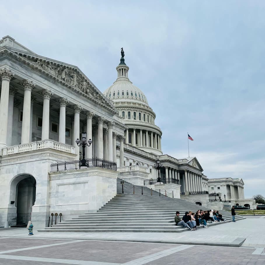 People seated on the steps of the U.S. Capitol, showcasing its architectural grandeur under a cloudy sky.