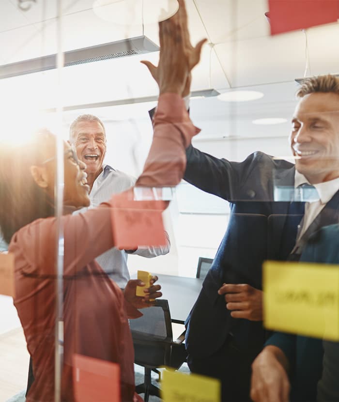 Colleagues celebrate in an office setting, sharing a high-five near a glass wall with colorful sticky notes.