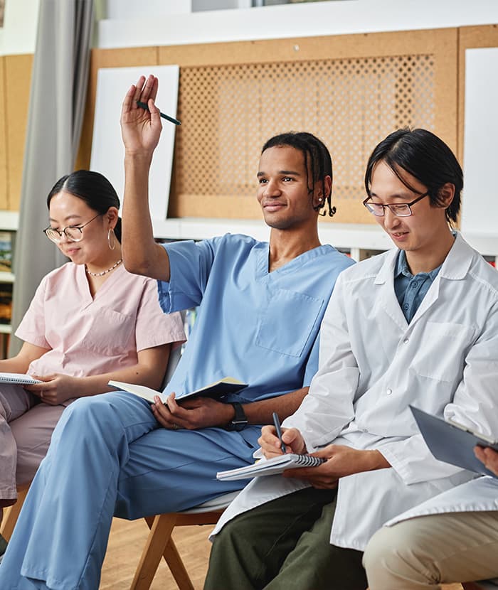 Group of diverse students in a classroom setting; one student raises their hand while others take notes.